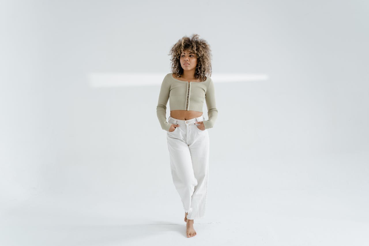 Stylish woman with afro hair and crop top posing barefoot in a bright studio shoot on white background.