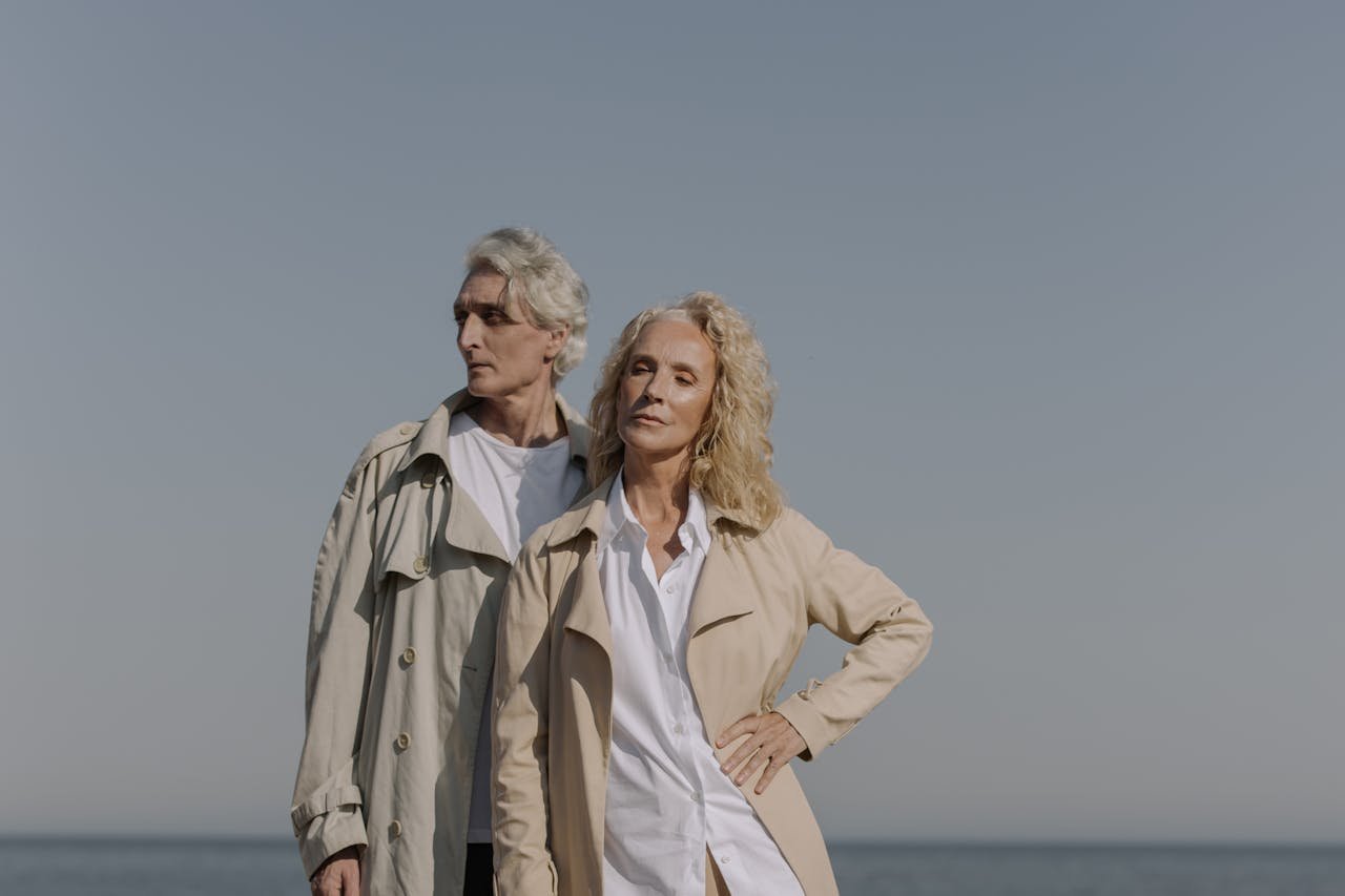 Stylish senior couple posing at the beach, under clear blue skies, wearing trench coats.