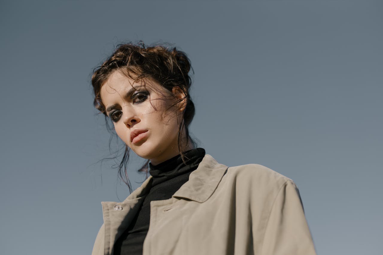 Elegant woman poses in a trench coat under clear skies, showcasing modern fashion.